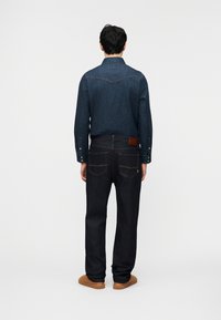 Άνδρας που φοράει σκούρο μπλε πουκάμισο από τζιν, σκούρο τζιν παντελόνι και καφέ παπούτσια, στέκεται με την πλάτη στραμμένη σε ένα απλό φωτεινό φόντο.