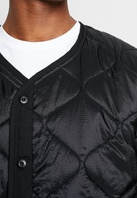 Musta tikattu toppatakki, jossa on nappikiinnitys, hienovarainen kiilto ja kankaan teksturoidulla timanttikuvioinnilla.