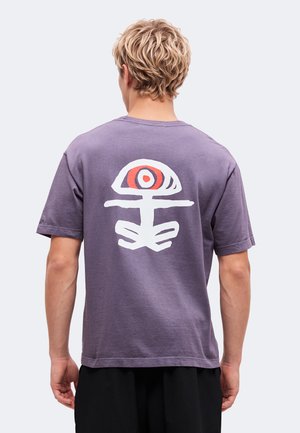 Blond person med kort hår iført en lilla t-shirt med et stort hvidt og rødt abstrakt øje- og figurdesign på bagsiden.