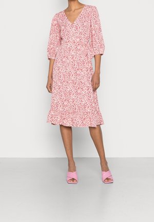 Femme portant une robe portefeuille rose à fleurs avec des manches trois-quarts et un ourlet à volants, associée à des sandales à talons ouvertes assorties roses.