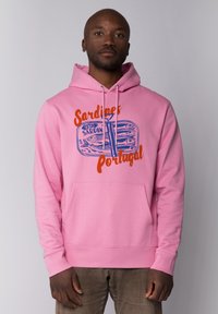Roze hoodie met een voorzak, voorzien van een grafisch ontwerp van een sardineblikje met blauwe en oranje tekst die "Sardines Portugal" zegt.