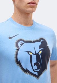 Κοντινό πλάνο ατόμου που φορά ανοιχτογαλάζιο πουκάμισο με μεγάλο σκούρο μπλε και κίτρινο θυμωμένο λογότυπο αρκούδας και μαύρο λογότυπο Nike στο στήθος.