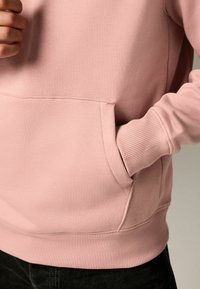Sudadera rosa claro con puños y dobladillo acanalados, que presenta un bolsillo delantero. El material parece suave y texturizado.