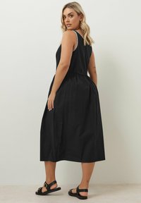 Svart ärmlös maxiklänning med rynkad midja, texturerad tyg, vit kant på axelsömmarna, ihop med svarta platta sandaler.