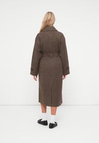 Brun herringbone frakke med beltestrop i taljen, bred krave og knaplukning ved ærmerne, med en knælang design og struktureret stof.