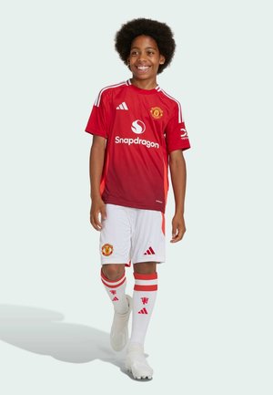 Rød fodboldtrøje med hvide shorts, præget af Manchester United-logoet og Snapdragon-branding. Hvide sokker med røde striber. Lys design.