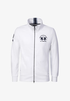 Valkoinen täyspitkä vetoketjullinen takki, jossa korkea kaulus, laivastonsininen polopelaajan logo rinnassa, raidallinen yksityiskohta kauluksen sisäpuolella ja merkki vasemmassa hihassa.