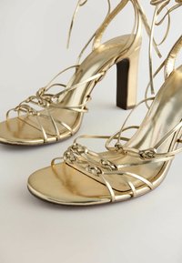 Sandales dorées à talons hauts avec brides, détails tressés et liens à nouer autour de la cheville sur un fond clair uni.