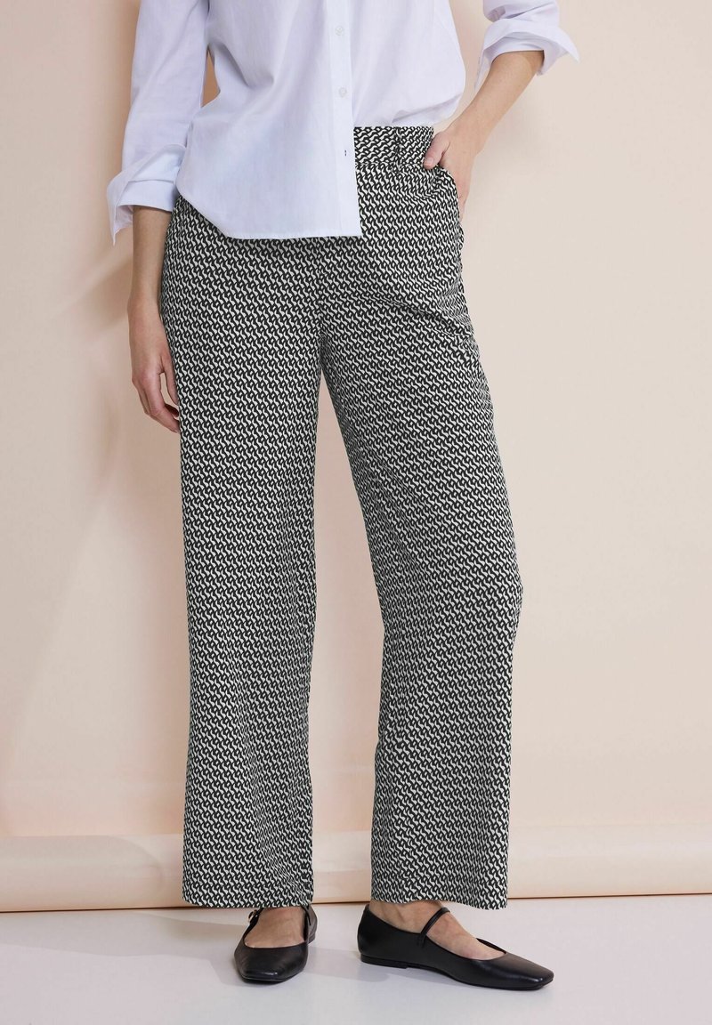 Personne portant un pantalon large à motifs noir et blanc, une chemise blanche boutonnée avec les manches retroussées, et des chaussures plates noires.