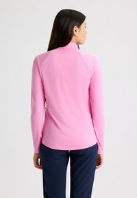 Rosa Langarmhemd aus glattem, dehnbarem Material mit steilem Kragen und Raglanärmeln. Getragen mit dunklen Marineblauen Hosen.