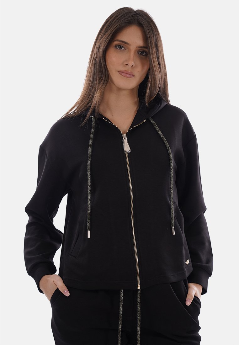 Femme portant un sweat à capuche noir zippé avec cordons, mains dans les poches, debout devant un fond clair uni, faisant face à l'avant.