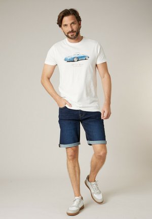 Homme portant un t-shirt blanc avec une impression de voiture décapotable bleue, un short en denim foncé retroussé et des baskets blanches, marchant vers l'avant.
