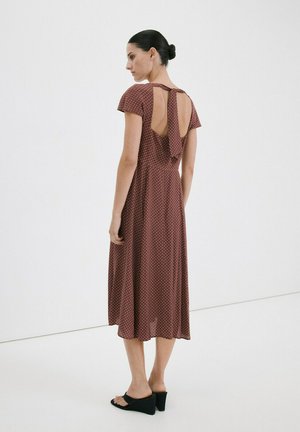 Mujer con cabello oscuro recogido en un moño, vistiendo un vestido midi marrón de manga corta con lunares, espalda descubierta y sandalias negras de tacón.