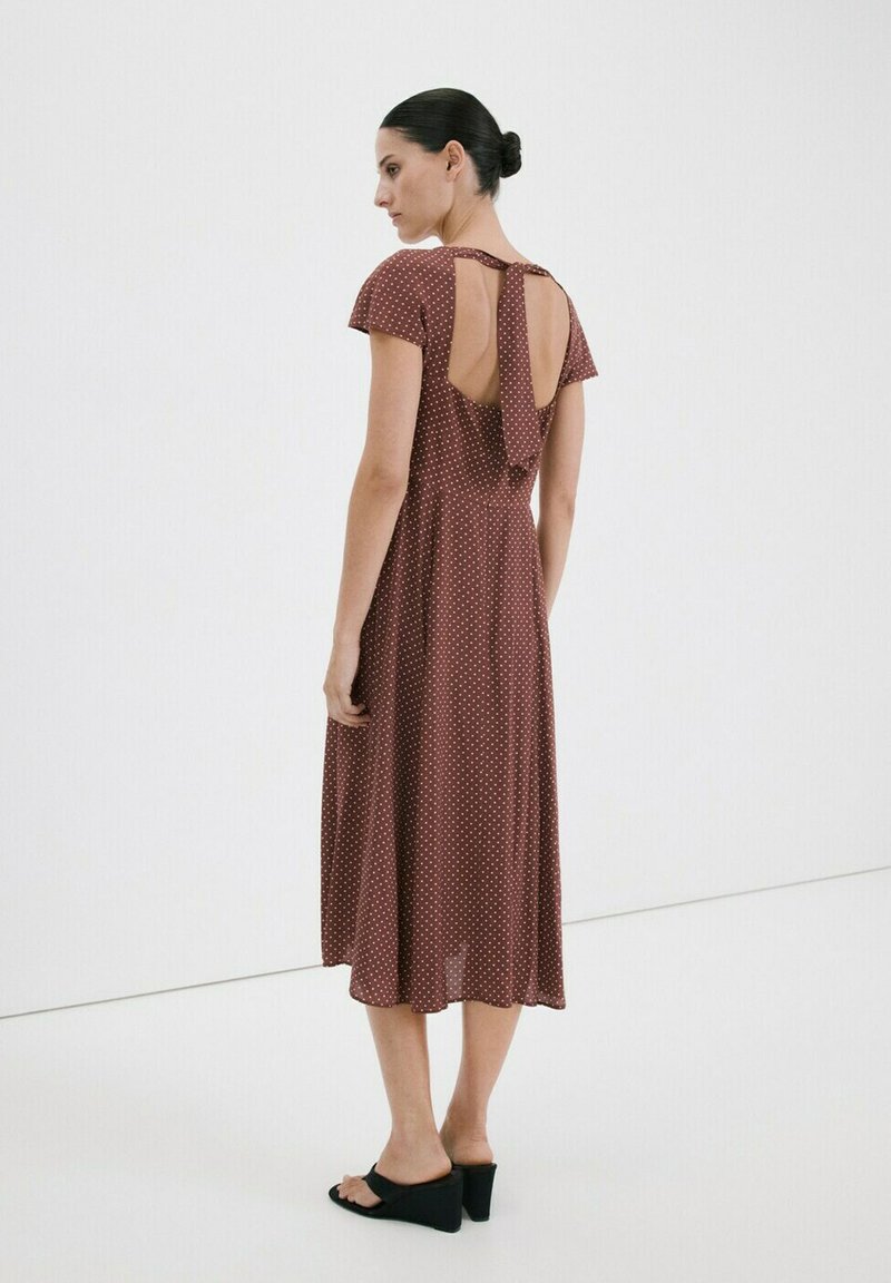 Žena s tmavými vlasmi v drdole, oblečená v hnedom midi šatách s krátkym rukávom a bodkami, s otvoreným chrbtom a čiernymi sandálmi na opätku.