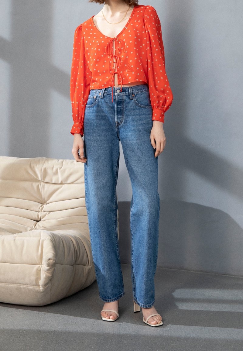 Femme portant un crop top rouge à manches longues avec des motifs blancs, un jean bleu taille haute et des sandales à talons beiges, debout à côté d'un canapé beige.