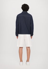 Mørkeblå denimjakke med spiss krage, synlig søm og knappede mansjetter, brukt over hvite shorts og sneakers.
