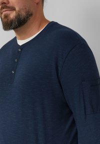 Navyblauw langarmshirt van een textuurstof met een ronde hals, voorzien van een drievoudige knoopsluiting en een zak op de linker mouw.