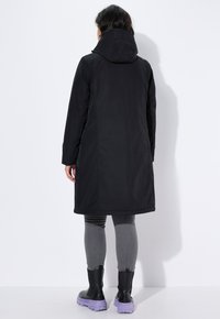 Mustat pitkät takki huppulla, jossa on sileä rakenne ja suora leikkaus. Käytetään harmaiden legginsien ja mustien vedenpitävien kenkien kanssa, joissa on violetit pohjat.