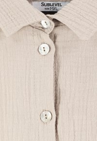 Beige button-up overhemd van textured stof. Heeft een puntige kraag en drie ronde knopen. Merkenlabel: Sublevel, maat M/L.