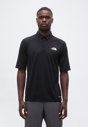 Uomo che indossa una polo nera a maniche corte con il logo The North Face sul petto sinistro e pantaloni grigi su sfondo semplice.
