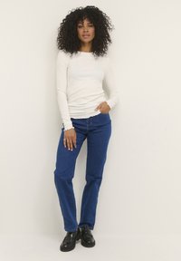 Top bianco a coste con maniche lunghe abbinato a jeans blu a gamba dritta e mocassini neri. Dettagli notevoli con polsini smerlati sul top.