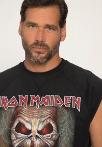 Černé tílko bez rukávů s velkým grafickým potiskem lebky s červenýma očima a plameny, s nápisem "Iron Maiden" v tučných písmenech.