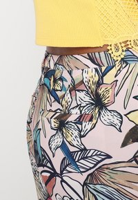 Żółty prążkowany crop top z koronkowymi akcentami, zestawiony z różowymi spodniami w kwiatowy wzór z dużymi liśćmi i kwiatami oraz boczną kieszenią.
