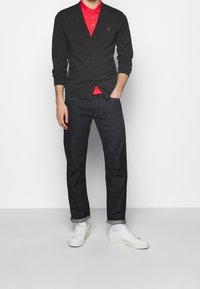 Uhlový kardigan s knoflíky, vrstvený přes červený polo, kombinovaný s tmavými džíny a bílými teniskami, s ohrnutými manžetami.