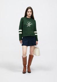 Green knitted sweater with white stripes, pleated navy skirt, brown knee-high boots, and a small white handbag.