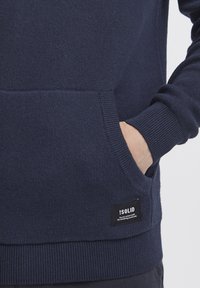 Sudadera azul marino de tejido de punto, con un bolsillo tipo canguro y puños acanalados. Se puede ver una etiqueta negra con texto blanco.