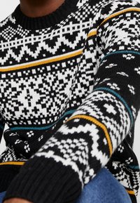 Pull en tricot noir et blanc à motifs avec des rayures jaunes et teal, doté d'un col rond et de poignets côtelés. Texture douce.