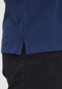 Sötétkék polo ing, textúrált anyagból, oldalsó hasítékkal és kontrasztos piros varrással az alján. Fekete nadrág látható alatta.