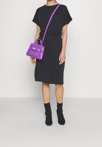 Lila, strukturierte Handtasche mit einer texturierten Oberfläche, kurzem Griff und verstellbarem Schulterriemen, kombiniert mit einem lockeren schwarzen Kleid und schwarzen Socken.