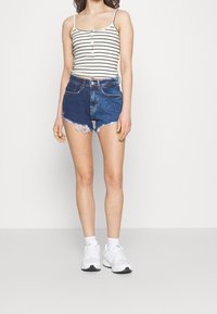 Gestreepte, mouwloze tanktop met knopen; blauwe denim korte broek met franje aan de onderkant; witte sneakers met grijze accenten; effen achtergrond.