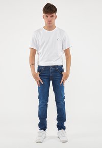 T-shirt bianca in cotone con un piccolo logo, abbinata a jeans denim blu scuro e sneakers bianche. Modella in piedi con le mani sui fianchi su uno sfondo bianco.