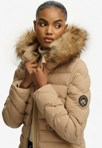 Beige dunjakke med pelsforet hætte, quiltet tekstur og lynlåslukning. Har logopatch på ærmet og høj krave.