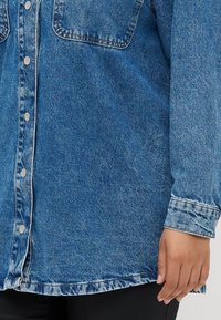 Blue denim jacket with snap buttons, chest pocket, and cuffed sleeves worn over black pants, hand relaxed at side.
