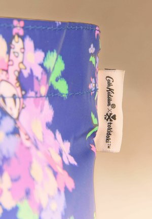 Zils audums ar krāsainu ziedu rakstu un baltu birku ar uzrakstu "Cath Kidston x tokidoki" uz bēša fona.