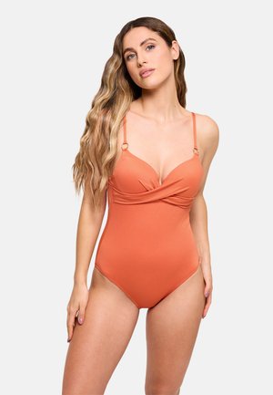 Mujer con el pelo largo y ondulado que lleva un traje de baño de una pieza texturizado en naranja con tirantes finos y un detalle frontal torcido.