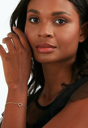 Goldenen Armreif mit Herzanhänger, der kleine klare Steine enthält. Ein Modell mit dunklen Haaren trägt ein transparentes schwarzes Oberteil, das den Schmuck an ihrem Handgelenk zeigt.