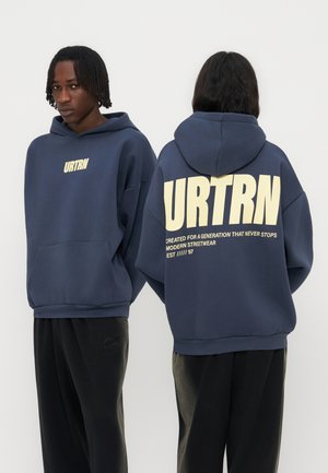Man draagt een donkerblauwe hoodie met het URTRN-logo aan de voorkant en grote URTRN-tekst met slogan op de achterkant, gecombineerd met een zwarte broek.