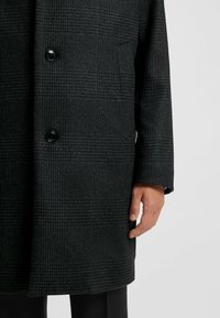 Cappotto doppiopetto in tessuto grigio scuro con motivo a quadri, caratterizzato da bottoni neri e un design a taglio dritto. Mano visibile.