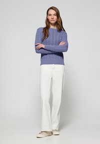 Ljuslila kabelstickad tröja, vita vida byxor och beige sneakers. Tröjan har ribbade ärmslut och fåll. Modellen står med armarna i kors.