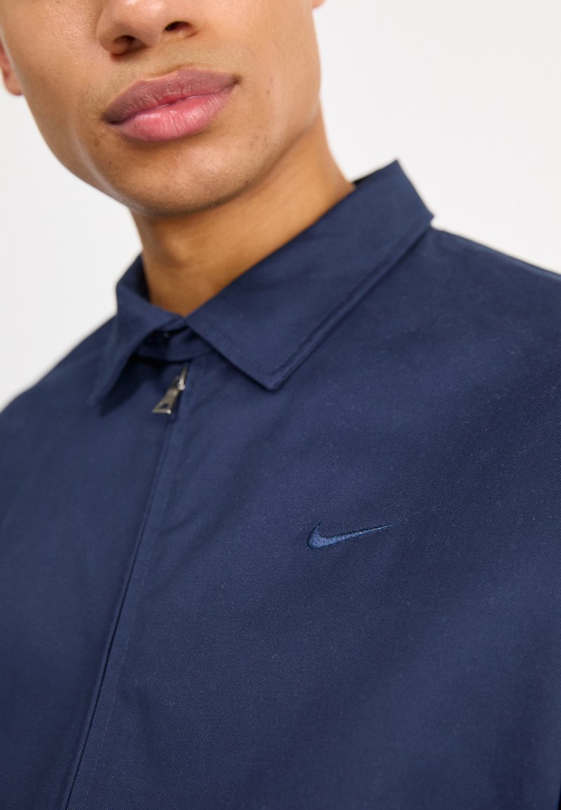 Veste bleu marine avec une texture lisse et un col pointu. Présente un petit logo Nike brodé sur le côté gauche. Fermeture zippée.