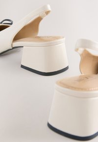 Paire de chaussures slingback écrues à talons blocs et semelles noires sur une surface claire.