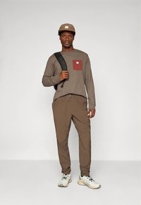 Langermet grå skjorte med en rød lomme, brune elastiske bukser og beige joggesko. Modellen bærer en svart ryggsekk og har på seg en beige caps.