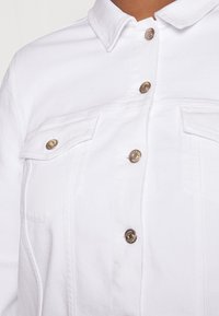 Gros plan sur une veste en denim blanche avec des boutons-pression en métal et des poches à rabat sur la poitrine, portée par une personne.