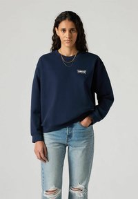 Granatowa bluza z miękkiego materiału, z małą naszywką z logo, okrągłym dekoltem i ściągaczami przy mankietach, połączona z wyblakłymi, poszarpanymi dżinsami.