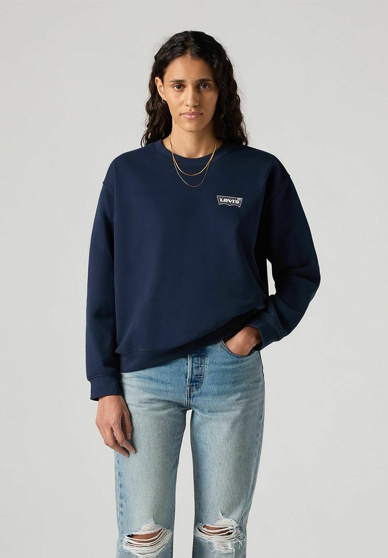 Granatowa bluza z miękkiego materiału, z małą naszywką z logo, okrągłym dekoltem i ściągaczami przy mankietach, połączona z wyblakłymi, poszarpanymi dżinsami.