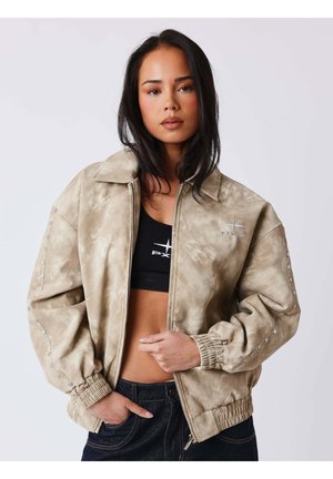 Jeune femme aux longs cheveux foncés, portant une veste beige par-dessus un crop top noir et un pantalon foncé, debout devant un fond clair uni.